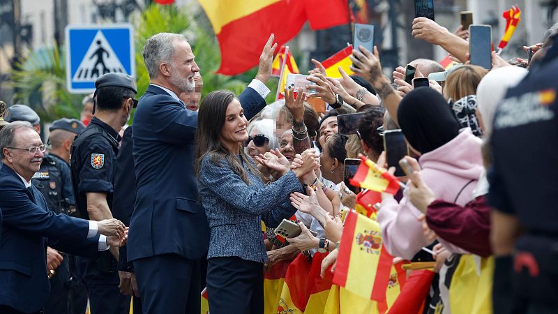 El Rey Felipe VI y la Reina Letizia saludan sonrientes a una multitud entusiasta que sostiene banderas de España y teléfonos móviles en un acto al aire libre.