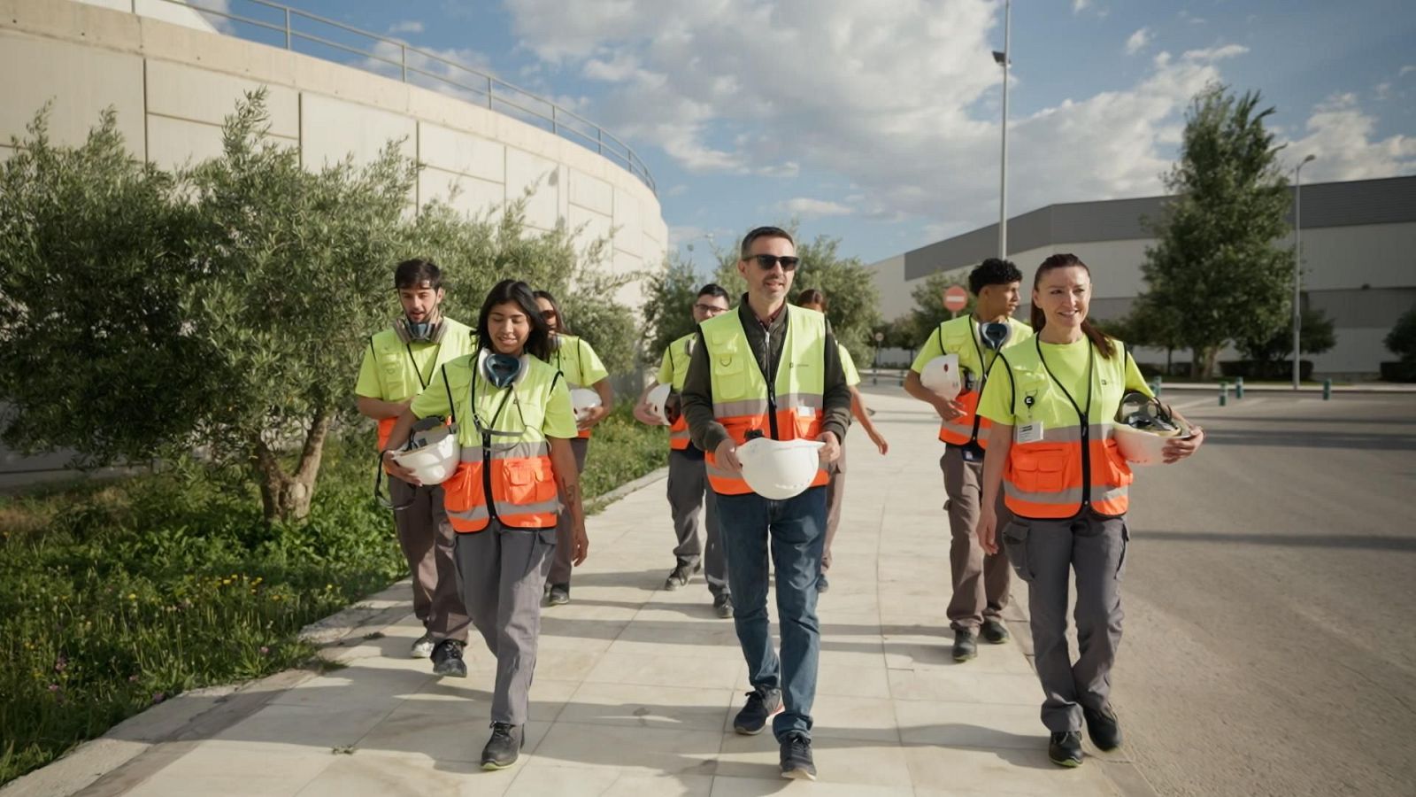 Grupo de personas con ropa de trabajo técnico y chalecos reflectantes camina por un entorno industrial portando cascos de seguridad.