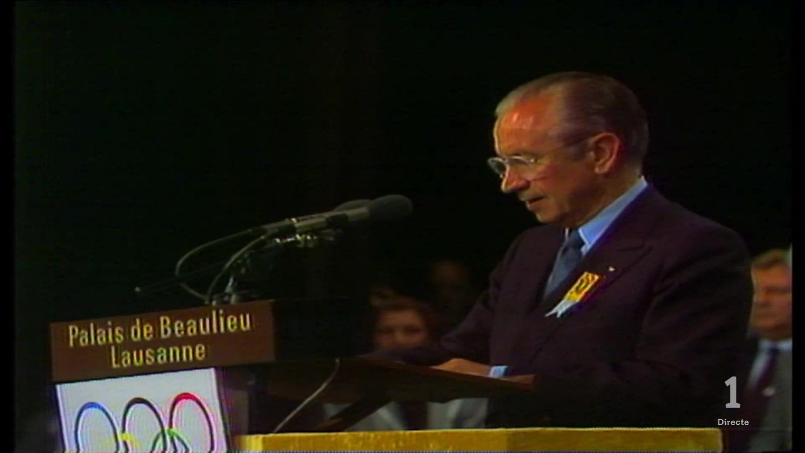Juan Antonio Samaranch de perfil tras un atril leyendo documentos durante la designación de Barcelona 1992, con micrófonos y el logotipo olímpico.