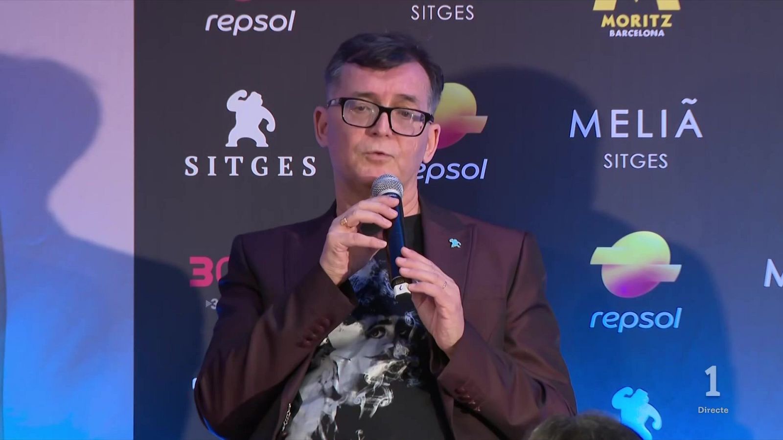 Ángel Sala, director del Festival de Sitges, participa en una rueda de prensa sosteniendo un micrófono inalámbrico frente a un panel de patrocinadores.