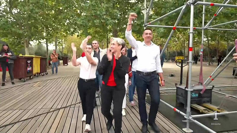 Tres personas caminan sonrientes y aplauden en una pasarela de madera al aire libre durante una retransmisión en directo.