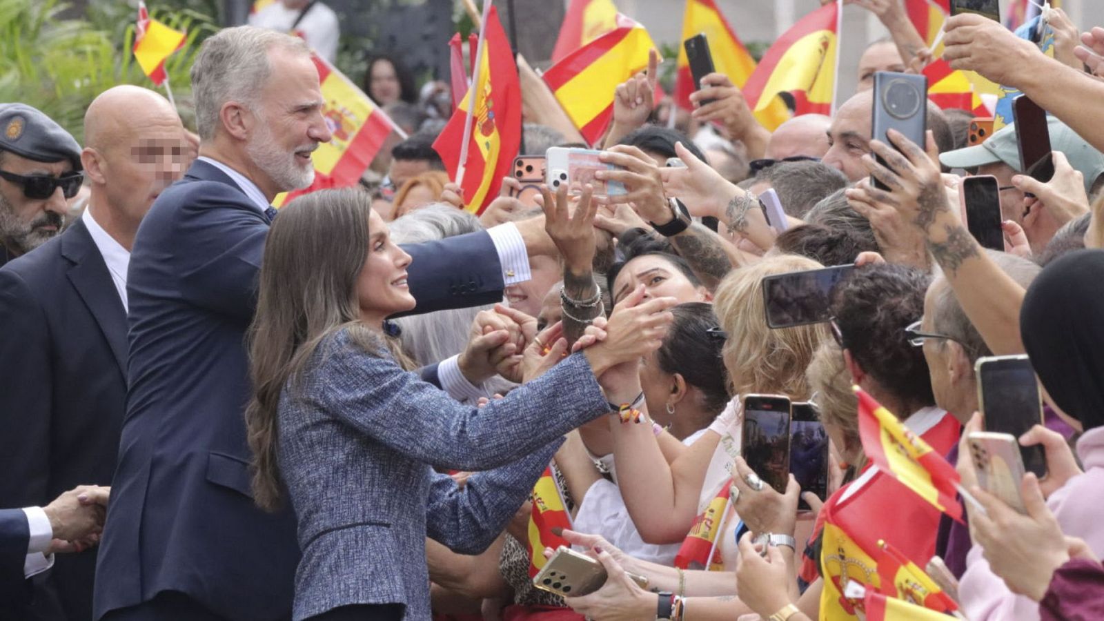 Los Reyes de España saludan cordialmente a una multitud entusiasta que alza teléfonos móviles y banderas.