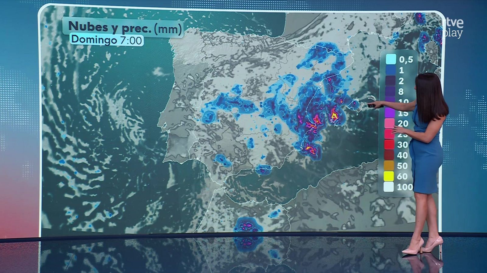 Presentadora en plató de informativos señala con un mando un mapa meteorológico de España con previsión de precipitaciones.