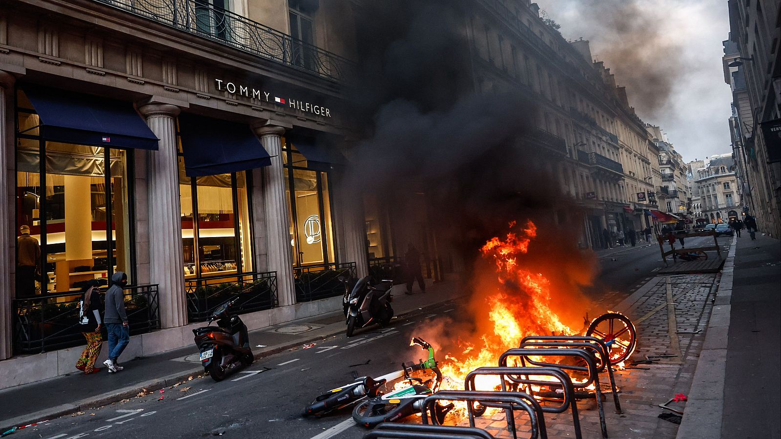 Calle céntrica de París durante disturbios con una hoguera en primer plano, una tienda de ropa y transeúntes en la acera.