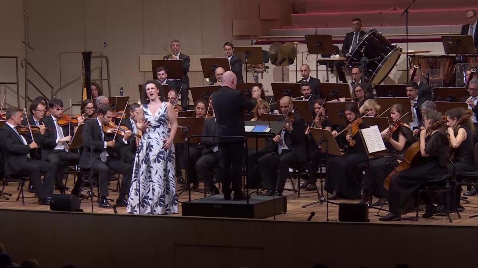 Cantante solista con vestido blanco y director de orquesta dirigiendo a los músicos en un escenario con iluminación cálida.