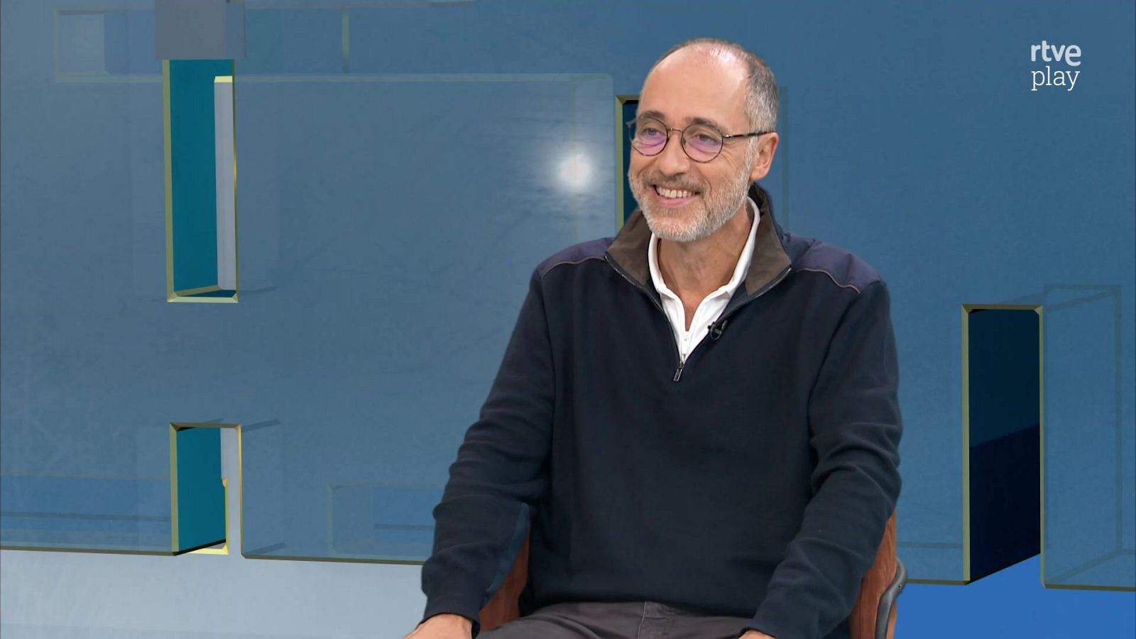 Hombre de mediana edad con gafas y barba canosa sonríe durante una entrevista en un plató con panel azul y logotipo de rtve play.