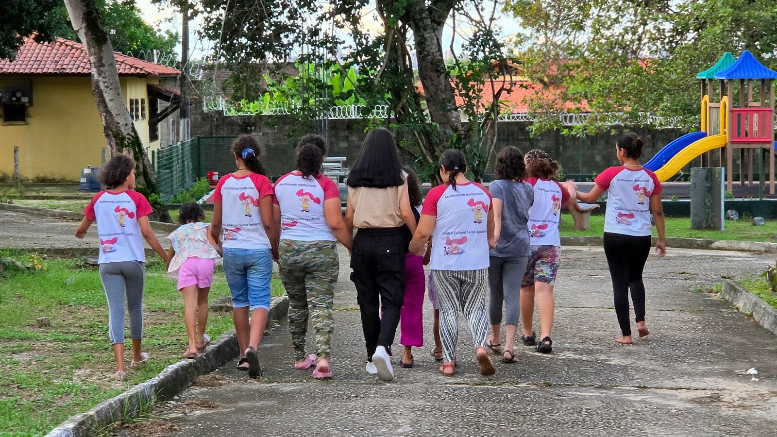 Un grupo de niñas y jóvenes caminan cogidas de la mano por un sendero exterior, vistiendo ropa informal y camisetas uniformadas frente a una zona con un parque infantil y árboles.