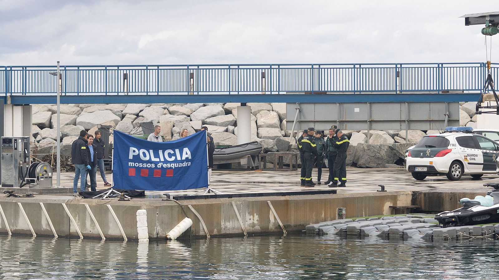 Operativo policial conjunto de los Mossos d'Esquadra y la Guardia Civil en un muelle portuario, con agentes, un coche patrulla y una lona de privacidad.