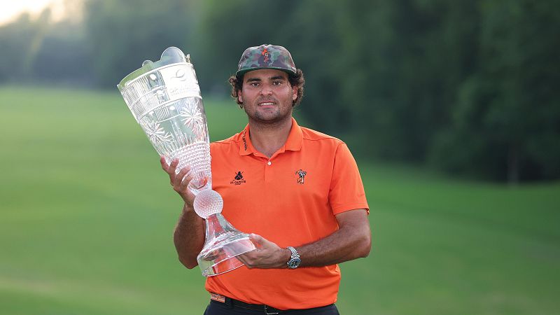 El golfista Ángel Hidalgo posa sonriente con un trofeo de cristal en un campo de golf, vistiendo un polo naranja y gorra de camuflaje.