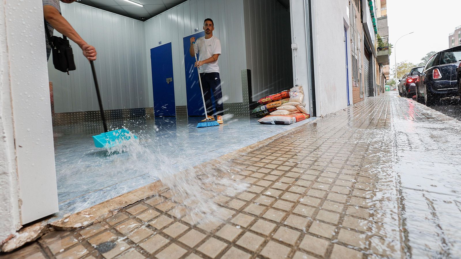 Dos personas achican agua tras una inundación en un local de trasteros, barriendo el suelo anegado hacia la acera.