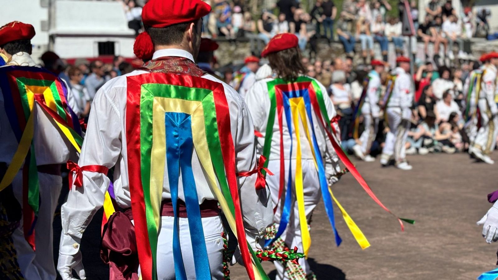 Danzantes tradicionales de espaldas con indumentaria blanca, boina roja y coloridas bandas en la espalda durante una celebración al aire libre con público al fondo.