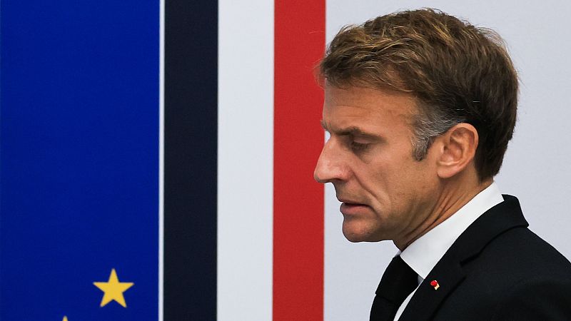 El presidente francés, Emmanuel Macron, aparece de perfil y con expresión seria, vistiendo traje formal y con las banderas de la UE y Francia de fondo.