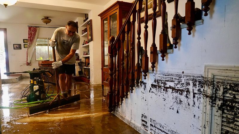 Un hombre con camiseta gris y guantes usa una rasqueta para achicar el agua y el lodo que inundan el interior de una vivienda familiar.