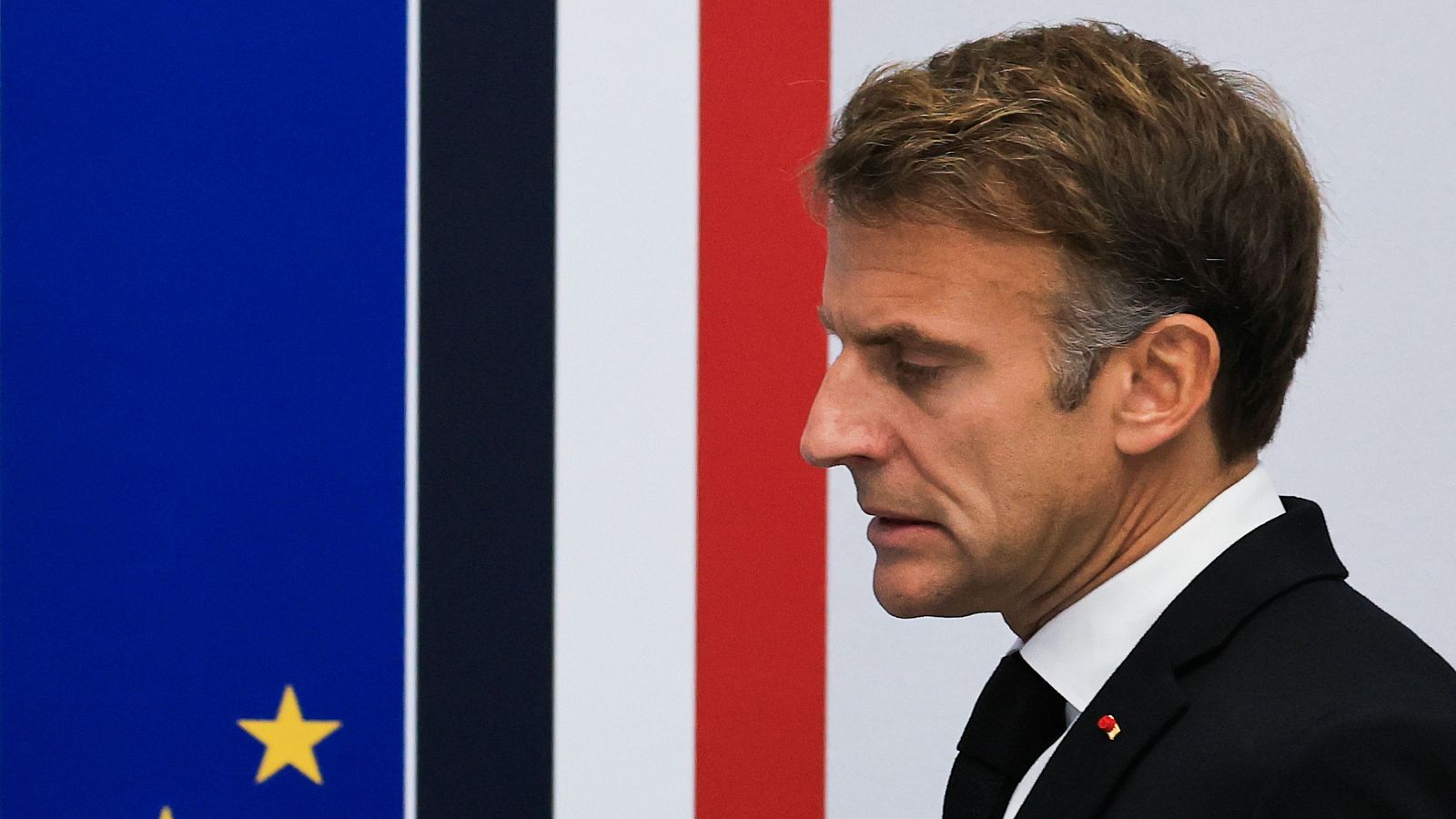 El presidente francés, Emmanuel Macron, aparece de perfil y con expresión seria, vistiendo traje formal y con las banderas de la UE y Francia de fondo.