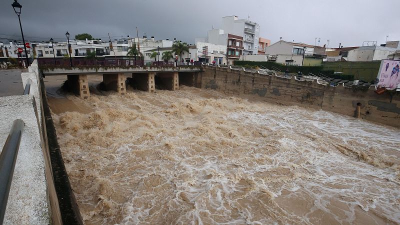 Violenta masa de agua marrón y turbia desborda el cauce canalizado de un río urbano bajo un puente y un cielo nublado.