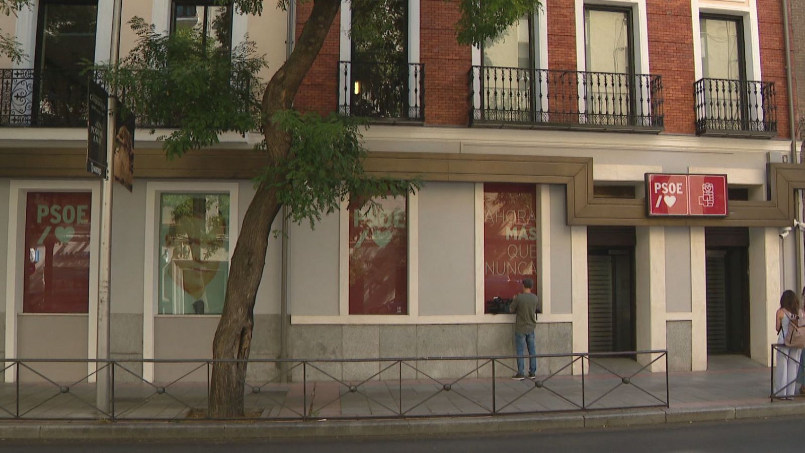 Fachada exterior de la sede de un partido político con carteles de campaña, ventanales de ladrillo visto y un cámara de televisión trabajando en la acera.