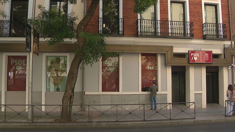 Fachada exterior de la sede de un partido político con carteles de campaña, ventanales de ladrillo visto y un cámara de televisión trabajando en la acera.