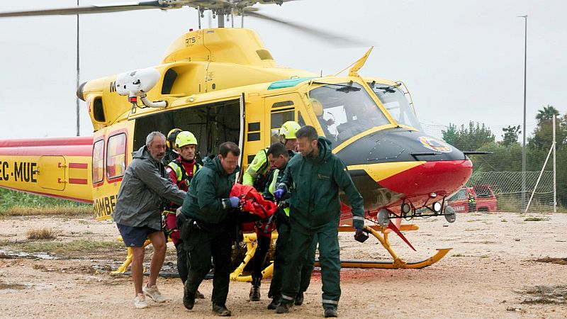 Equipo de emergencias y Guardia Civil transportan una camilla roja en un terreno embarrado junto a un helicóptero amarillo y rojo.