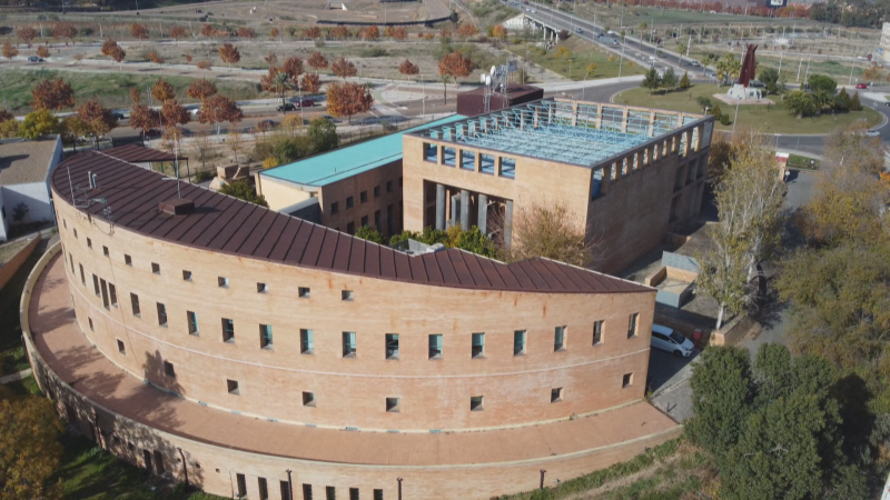 Vista aérea diurna de las instalaciones de RTVE en Mérida, con un edificio semicircular de ladrillo visto, tejado rojizo y un bloque anexo con celosía metálica azul y antenas.