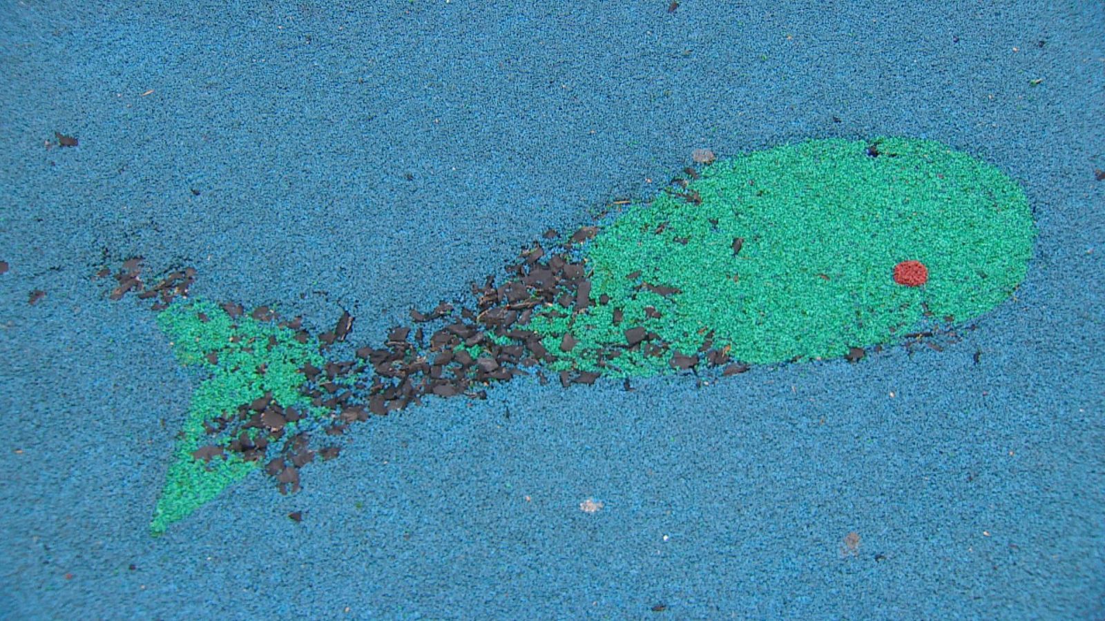 Suelo de caucho continuo de un parque infantil con un diseño de pez verde sobre fondo azul, que muestra grietas y desprendimientos.