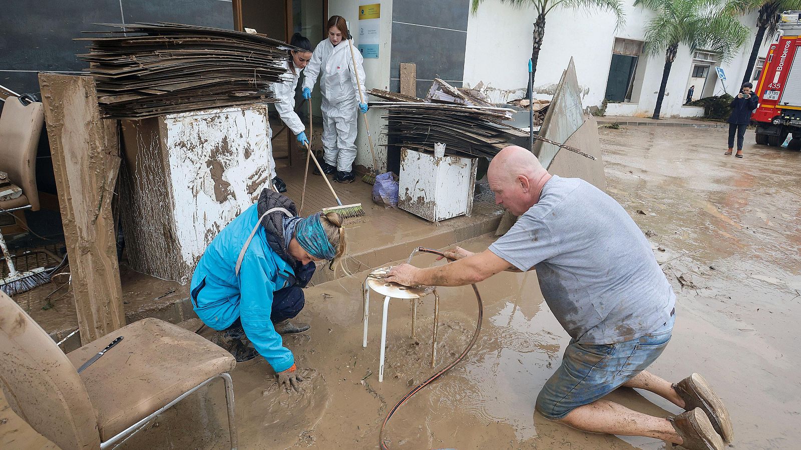 Personas limpiando con mangueras y herramientas el lodo acumulado en la calle y la entrada de un edificio tras una inundación.