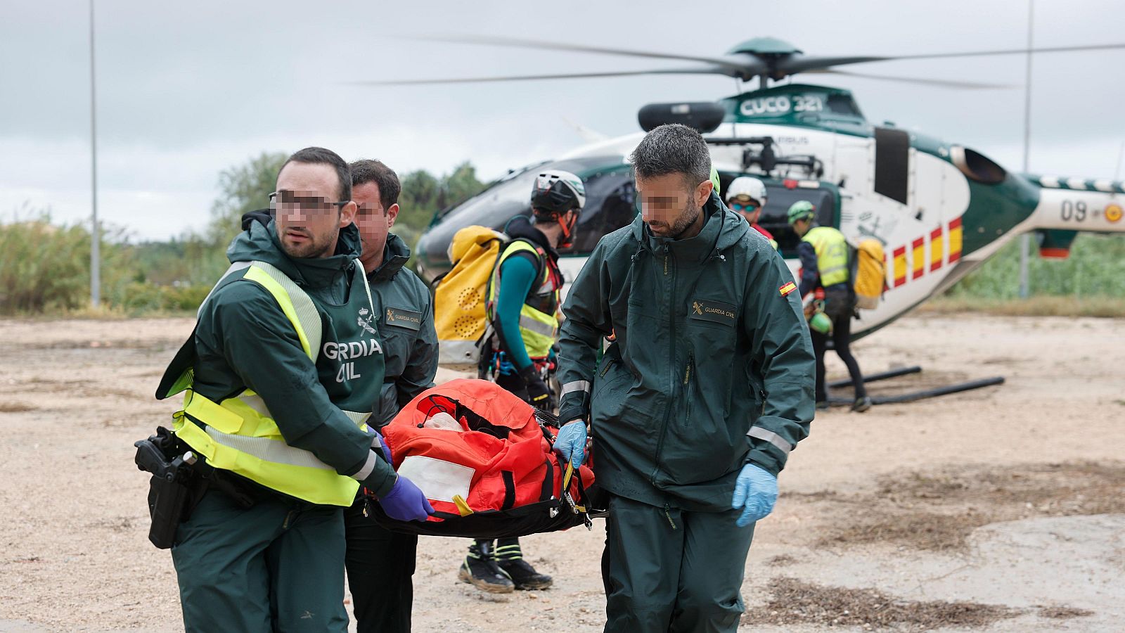 Dos agentes de la Guardia Civil con anorak verde y chaleco reflectante transportan una camilla de rescate roja en un terreno natural bajo un cielo nublado.