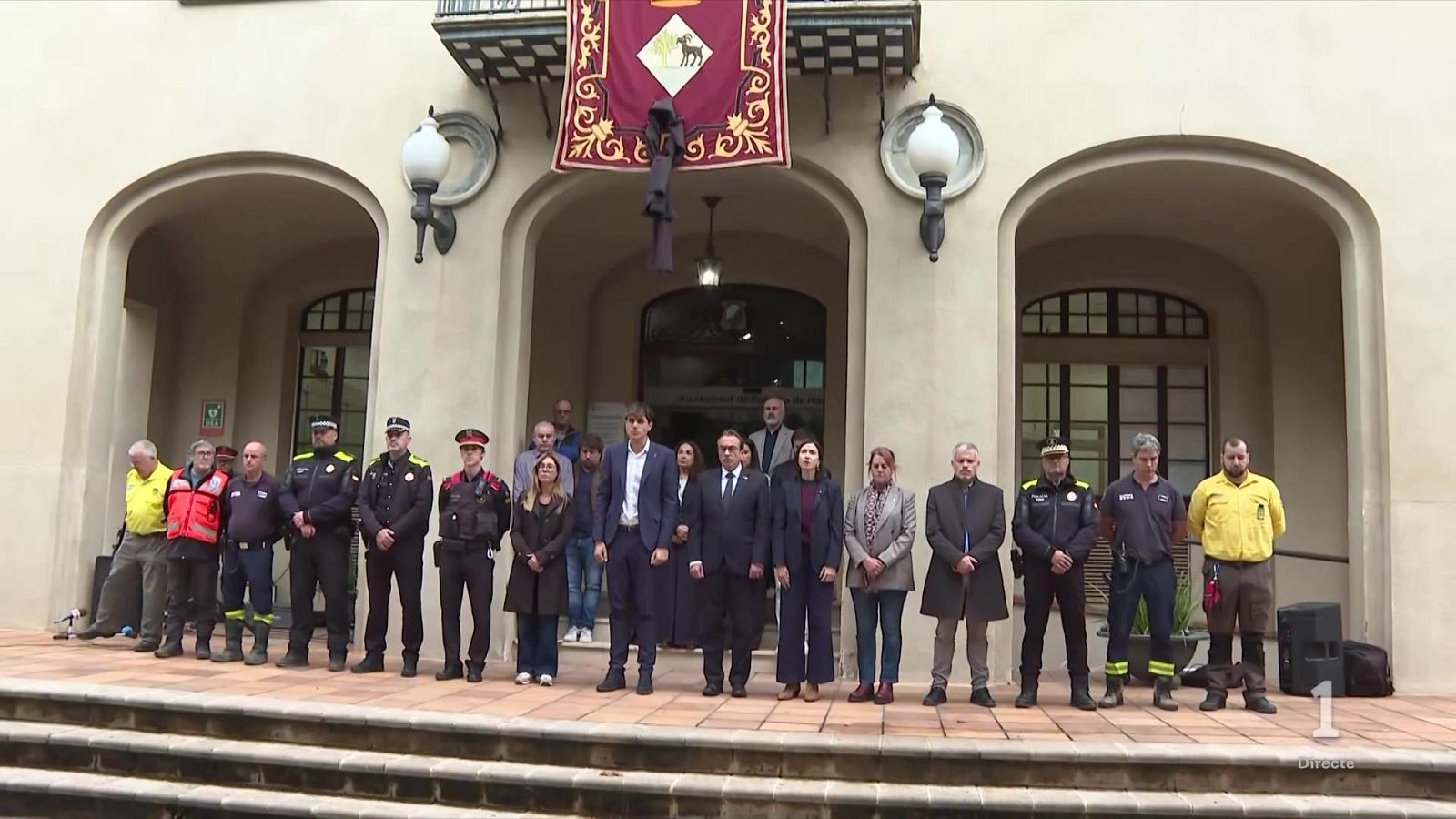 Acto institucional solemne con autoridades, policías y servicios de emergencia guardando un minuto de silencio frente a un edificio municipal con un crespón negro.