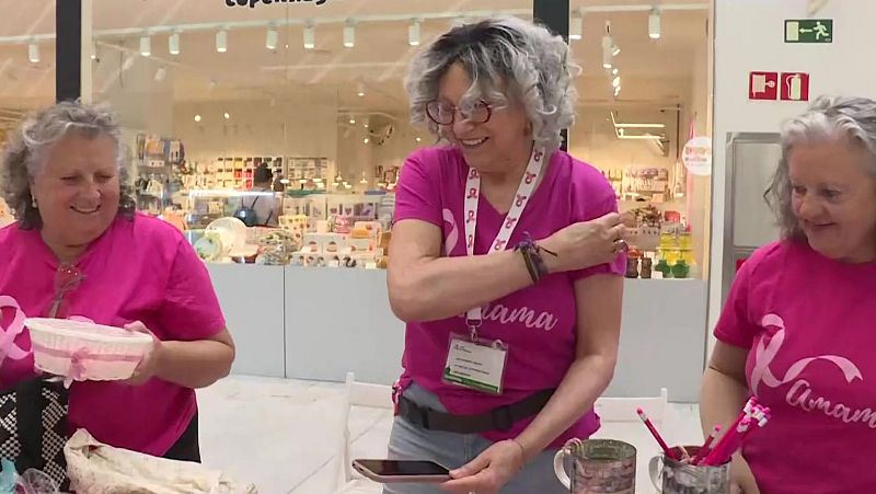 Tres voluntarias con camisetas rosas y tarjetas identificativas atienden una mesa informativa contra el cáncer de mama en un centro comercial.