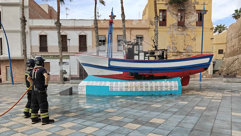 Dos bomberos apagan los restos humeantes de un monumento con forma de barco pesquero calcinado en una plaza urbana rodeada de edificios.