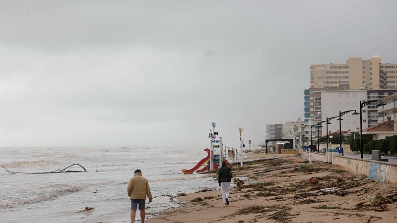 Playa urbana tras un temporal marítimo con fuerte oleaje, arena erosionada con residuos, zona de juegos infantil y paseo marítimo con edificios al fondo.