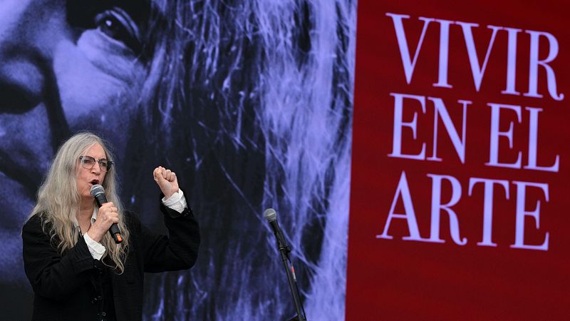 La cantante Patti Smith actúa en un escenario con un micrófono y chaqueta oscura, frente a un telón con su rostro y el lema «VIVIR EN EL ARTE».