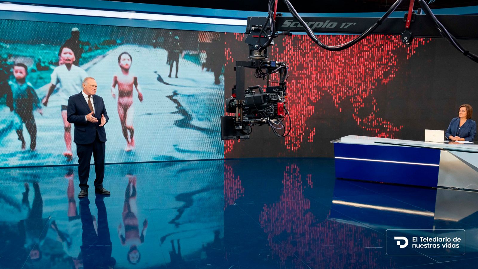 Plató de informativos con pantalla LED curva, periodista de pie, presentadora sentada en mesa y cámara de grúa colgada.