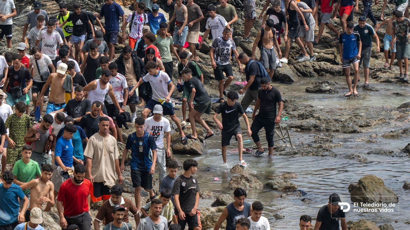 Multitud de jóvenes avanzan a pie por una costa rocosa y resbaladiza junto al mar en una crisis migratoria