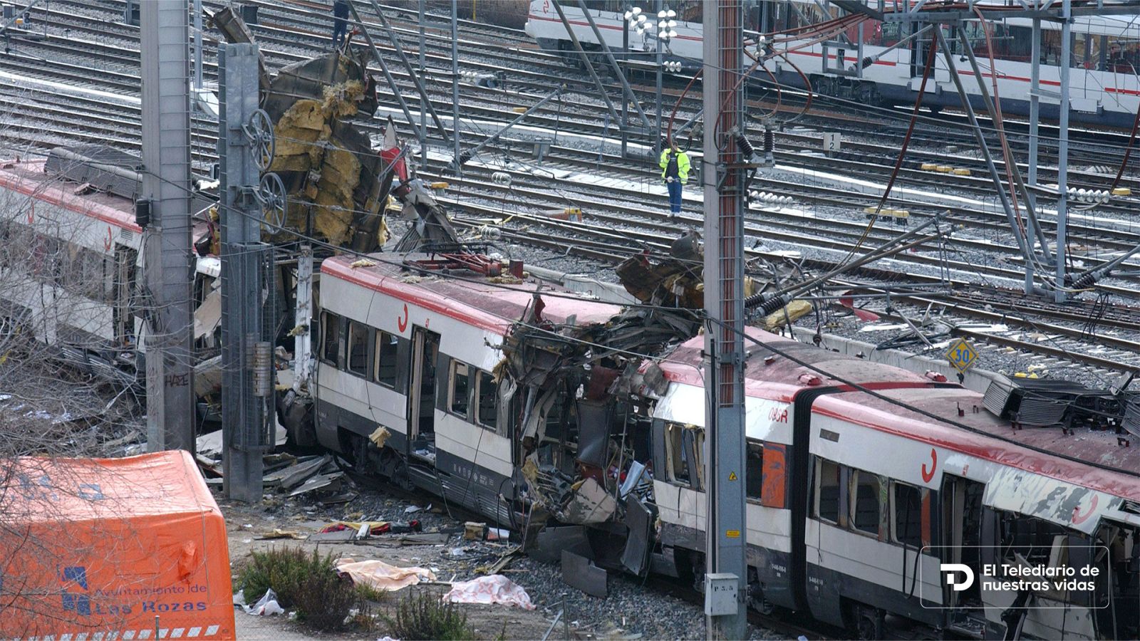 Vista cenital de un tren de Cercanías de Renfe gravemente destruido tras una explosión en las vías de Madrid, con una carpa de emergencias naranja en el suelo.
