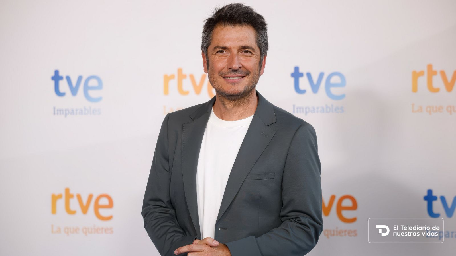Plano medio del periodista Carlos del Amor posando sonriente ante un photocall de RTVE con americana gris y camiseta blanca.