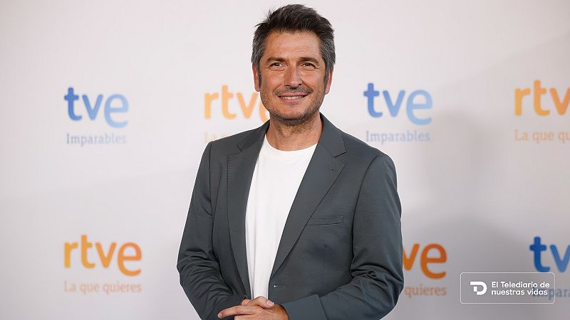 Plano medio del periodista Carlos del Amor posando sonriente ante un photocall de RTVE con americana gris y camiseta blanca.