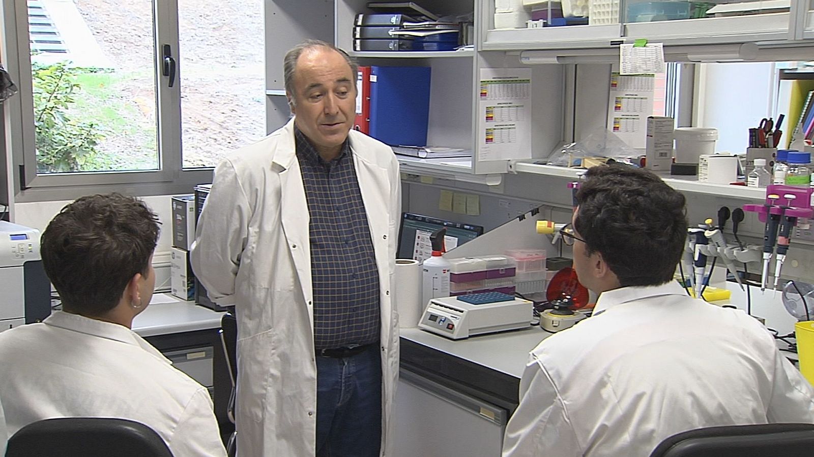 Tres investigadores con bata blanca debaten en un laboratorio junto a una poyata con instrumental científico, micropipetas y tubos de ensayo.