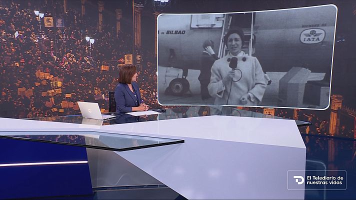 Plató de informativos con una presentadora ante una mesa blanca y moderna, mirando una pantalla flotante que muestra una reportera histórica junto a un avión.