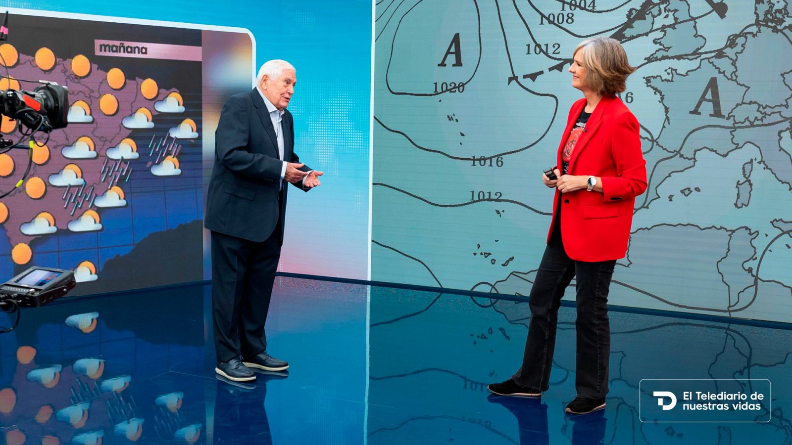 Dos presentadores del tiempo conversan en un plató de televisión junto a pantallas con mapas meteorológicos modernos e históricos.