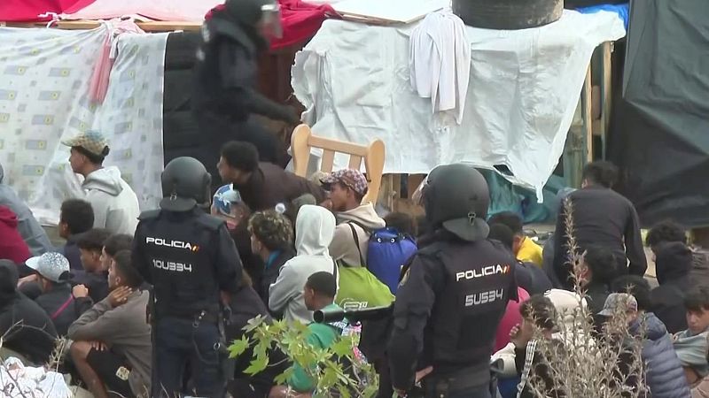Agentes antidisturbios de la Policía Nacional custodian a un grupo de jóvenes sentados en el suelo junto a refugios improvisados.