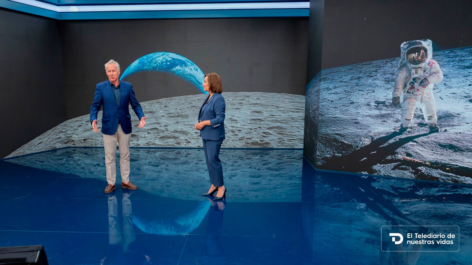 Plató de informativos con suelo azul reflectante y pantallas gigantes que muestran la superficie lunar y un astronauta.