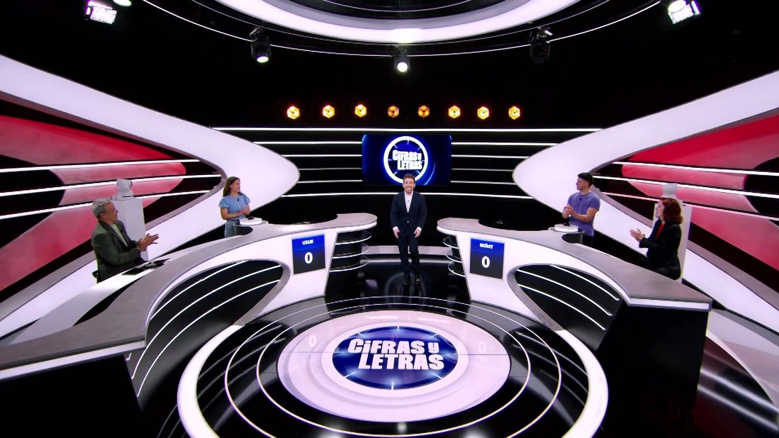 Plató de televisión futurista con el presentador y dos expertos acompañando a dos concursantes junto a sus atriles con marcadores a cero.