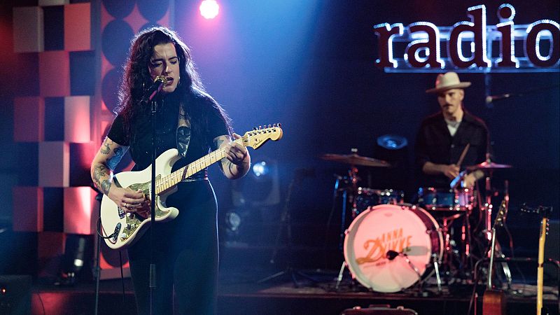 La cantante Anna Dukke interpreta con pasi�n una guitarra el�ctrica junto a su baterista en un plat� iluminado con luces azules y rojas.