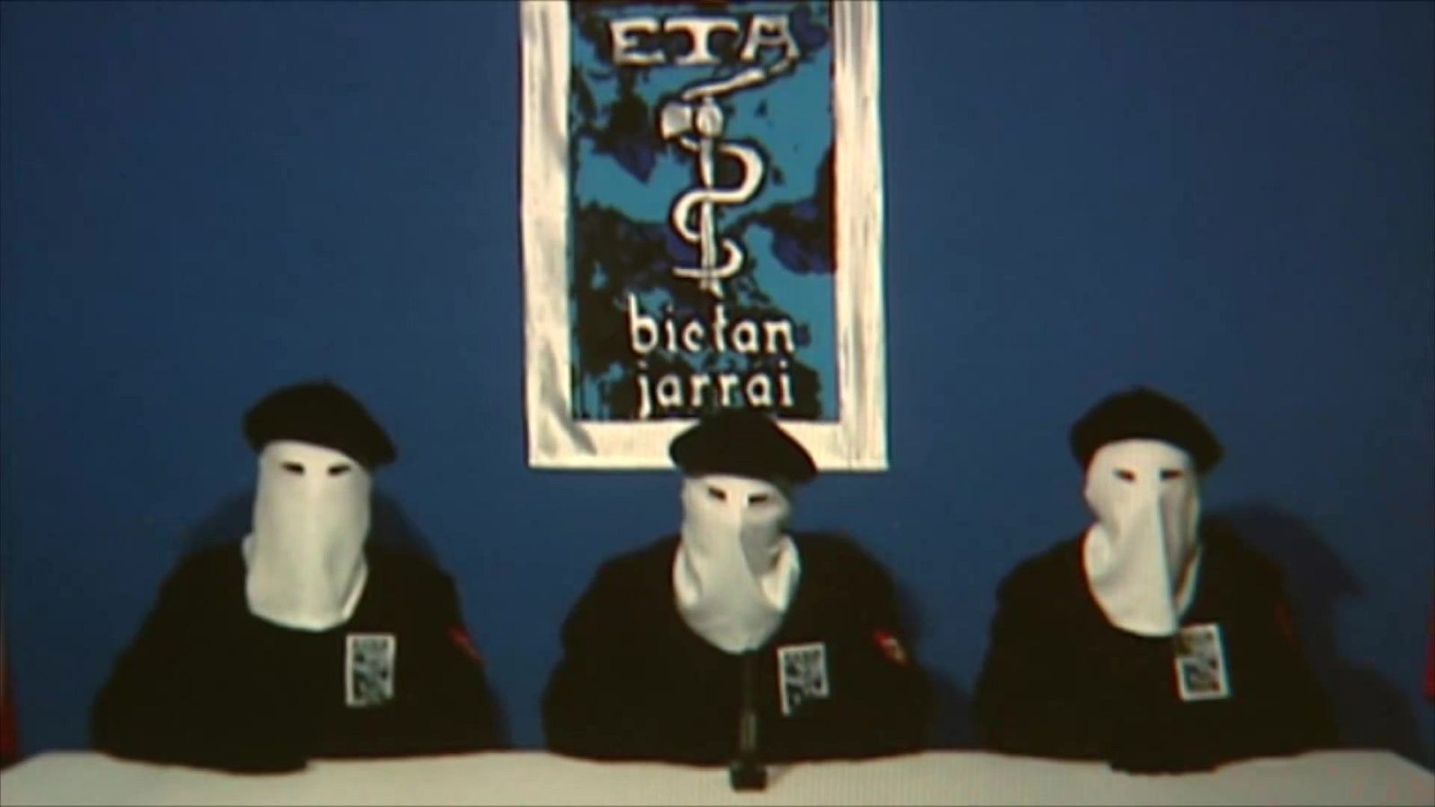 Tres enmascarados con capuchas blancas y boinas negras sentados ante una mesa con el anagrama de ETA de fondo.