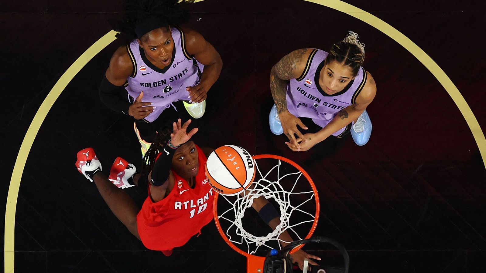 Vista cenital de una jugada de baloncesto con una atacante roja de Atlanta Dream y dos defensoras de Golden State bajo el aro.