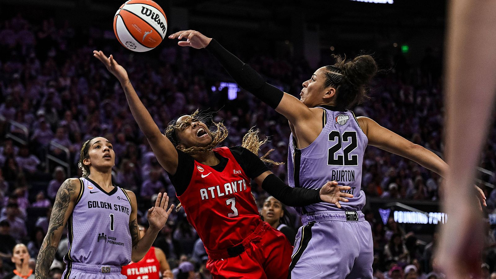 Intensa disputa aérea por el balón entre dos jugadoras de baloncesto con uniformes rojo y violeta en un pabellón lleno de espectadores.