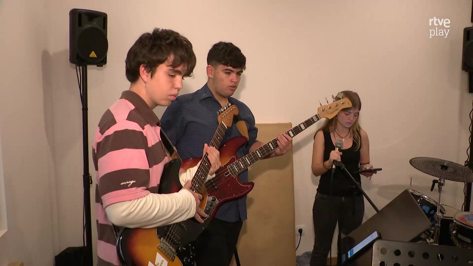 Tres jóvenes ensayan en un local de música equipados con guitarra eléctrica, bajo, micrófono y otros instrumentos.
