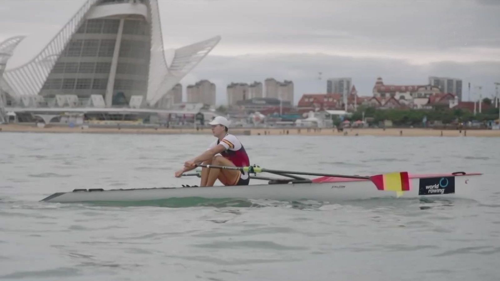 Un remero español con uniforme nacional navega en un bote individual sobre aguas tranquilas frente a la costa urbana.