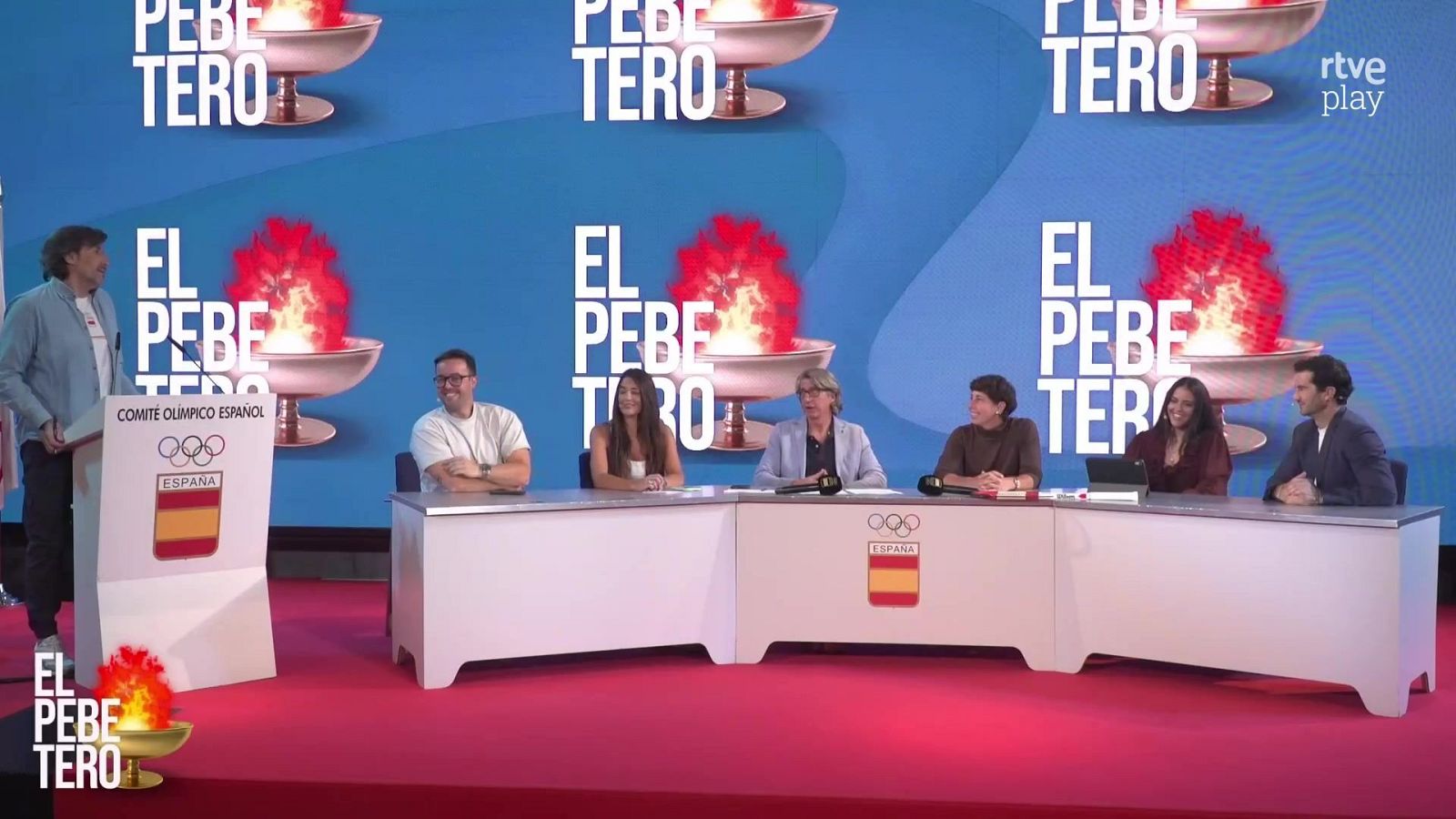 Plató de televisión de 'El pebetero' con un presentador en pie junto a un atril y seis tertulianos sentados en una mesa curvada frente a un gran videowall azul.