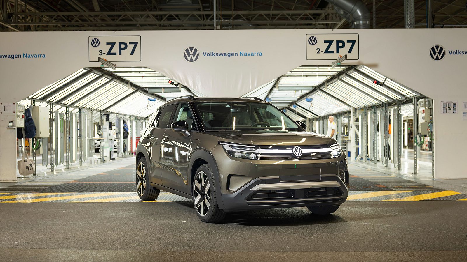 Vehículo crossover compacto de color bronce de Volkswagen en la salida de un túnel de inspección de una fábrica, con suelo de hormigón y marcas de seguridad amarillas.